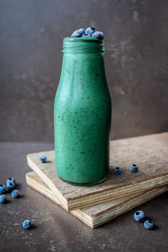 Glowing Green Spirulina Smoothie in a glass from the side with blueberries on top and on the sides