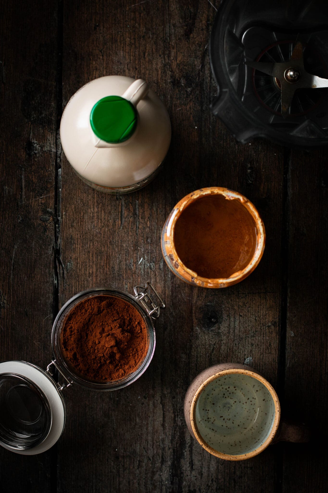 Maple, almond butter, cocoa powder, a mug and a blender seen from the top.