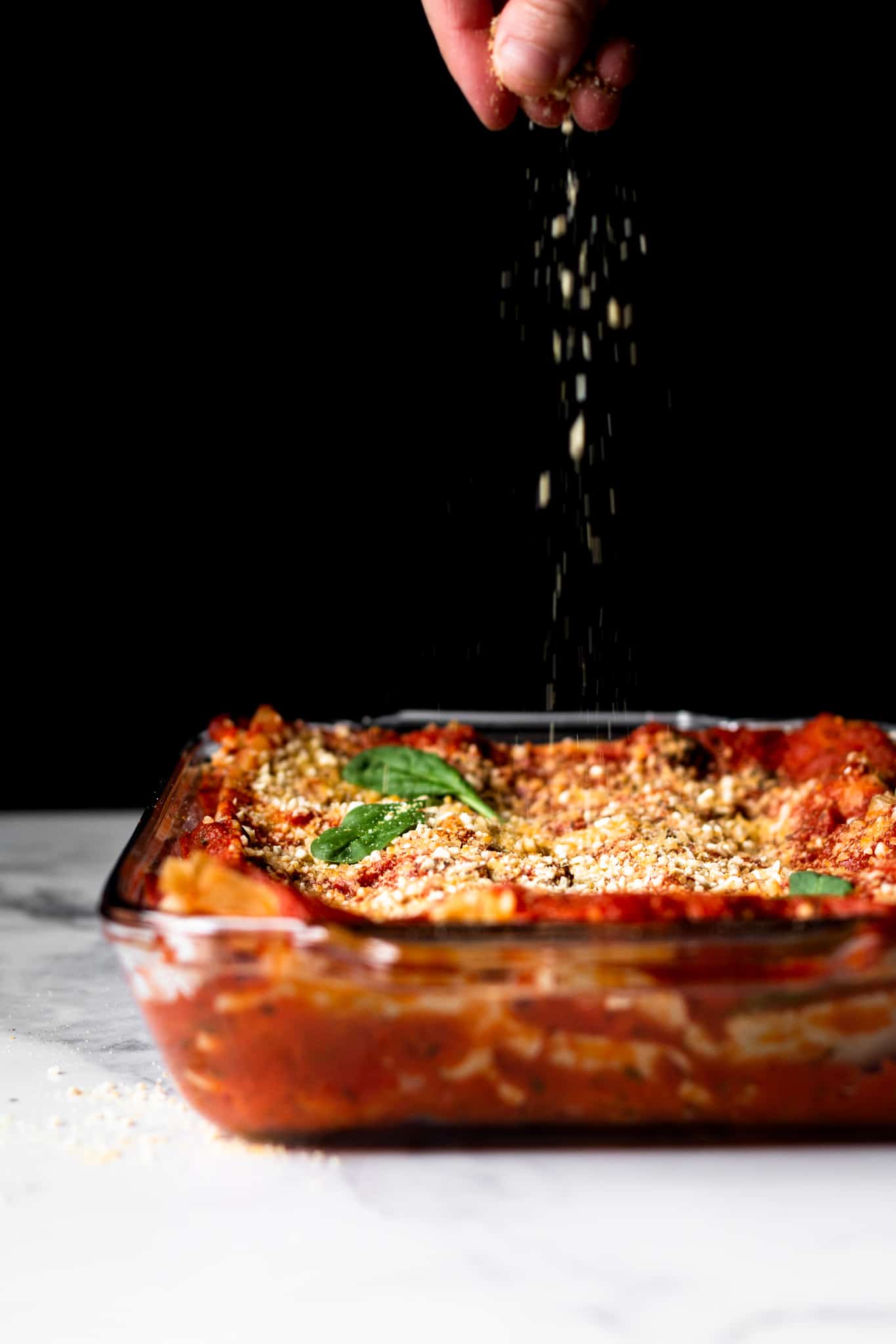 hand sprinkling vegan parmesan on lasagna