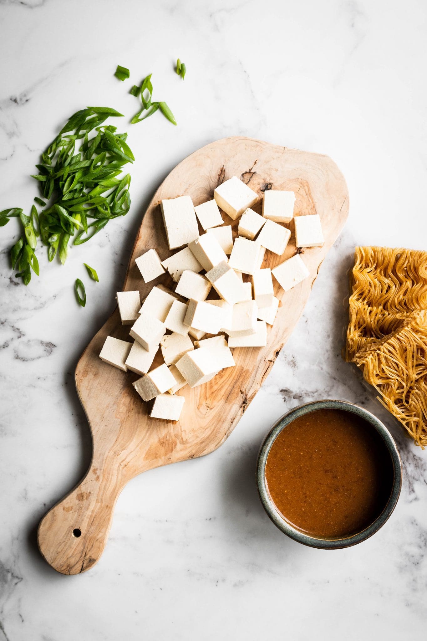 cubed tofu on a cutting board with scallions, ramen noodles and sauce