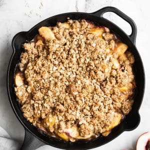 vegan peach crisp in a cast-iron skillet with a sliced peach on the side