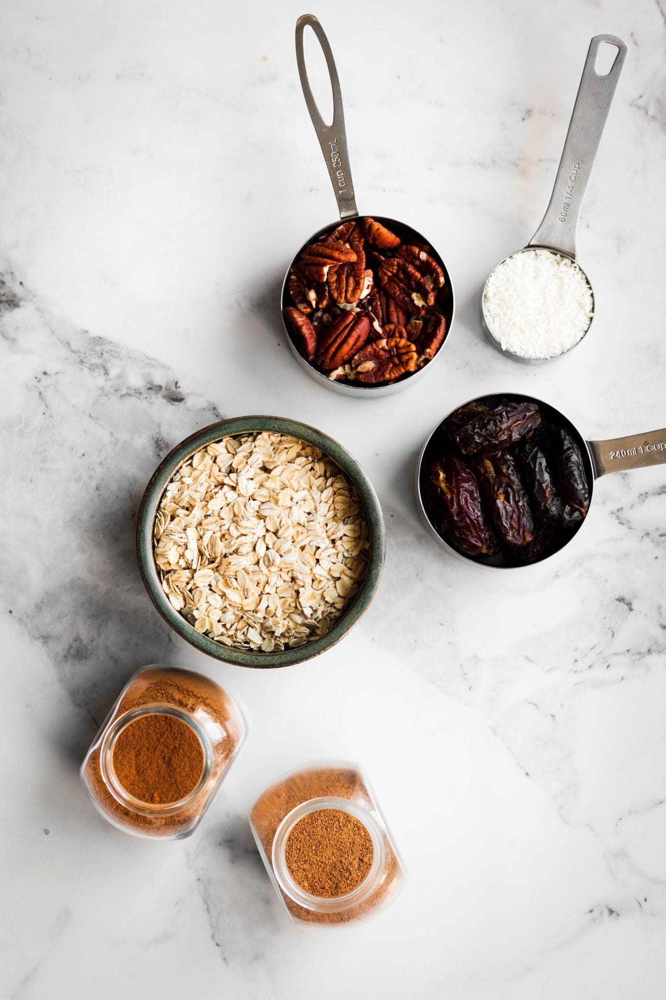 pecans, coconut, oats, dates and spices in measuring cups and bowls seen from the top