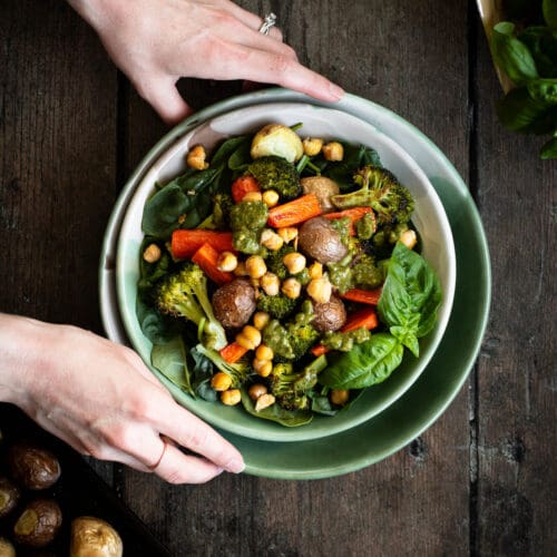 hands holding a bowl of roasted vegetables with almond pesto seen from the top