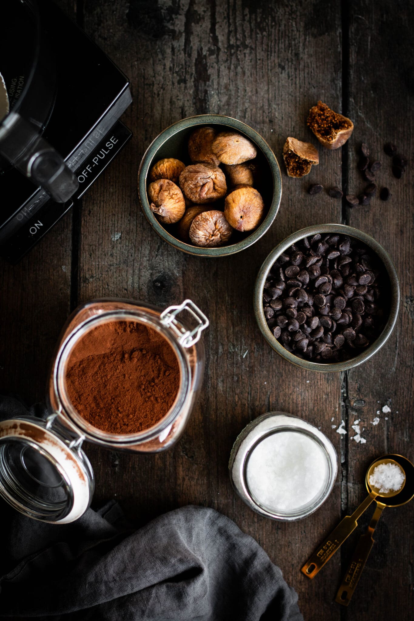 Dried figs, chocolate chips, salt and cocoa powder in bowls seen from the top.